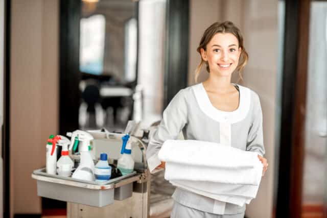 Young-woman-chambermaid holding a towel standing with maid cart full of cleaning stuff in the hotel corridor