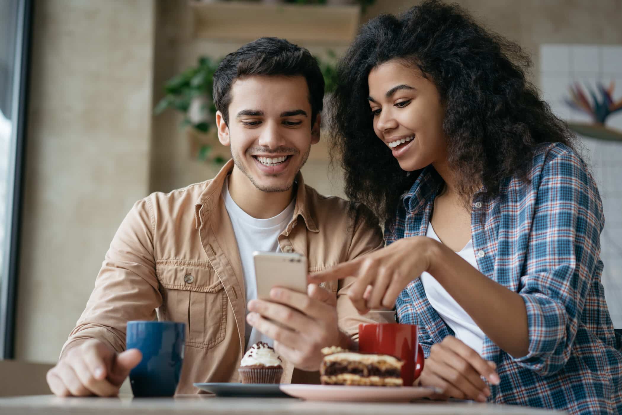 Happy couple using mobile phone application for online shopping. Emotional friends communication, laughing, looking at digital screen, sitting together in cafe