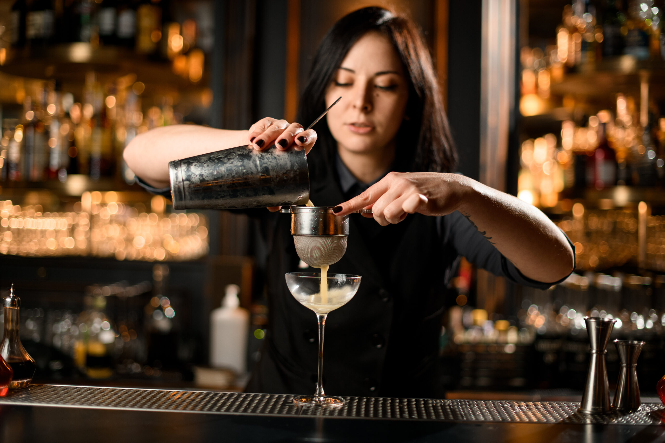 female bartender makes cocktail drink with a steel sieve