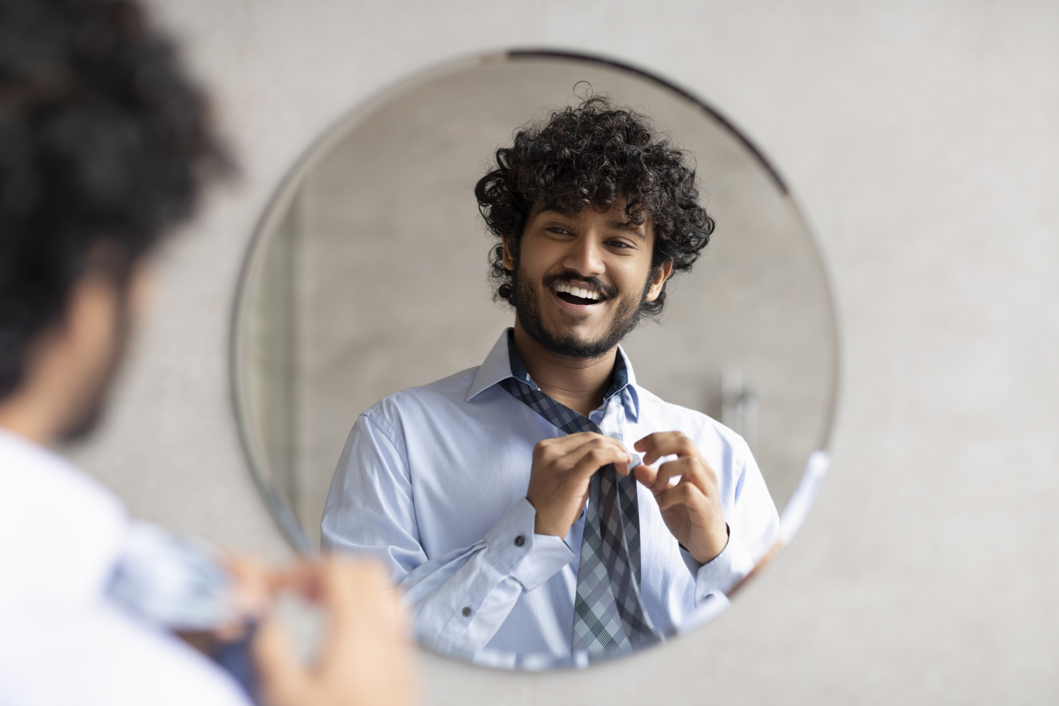 Happy indian man putting on necktie while looking in the mirror and smiling at his reflection, standing in modern bathroom interior, free space