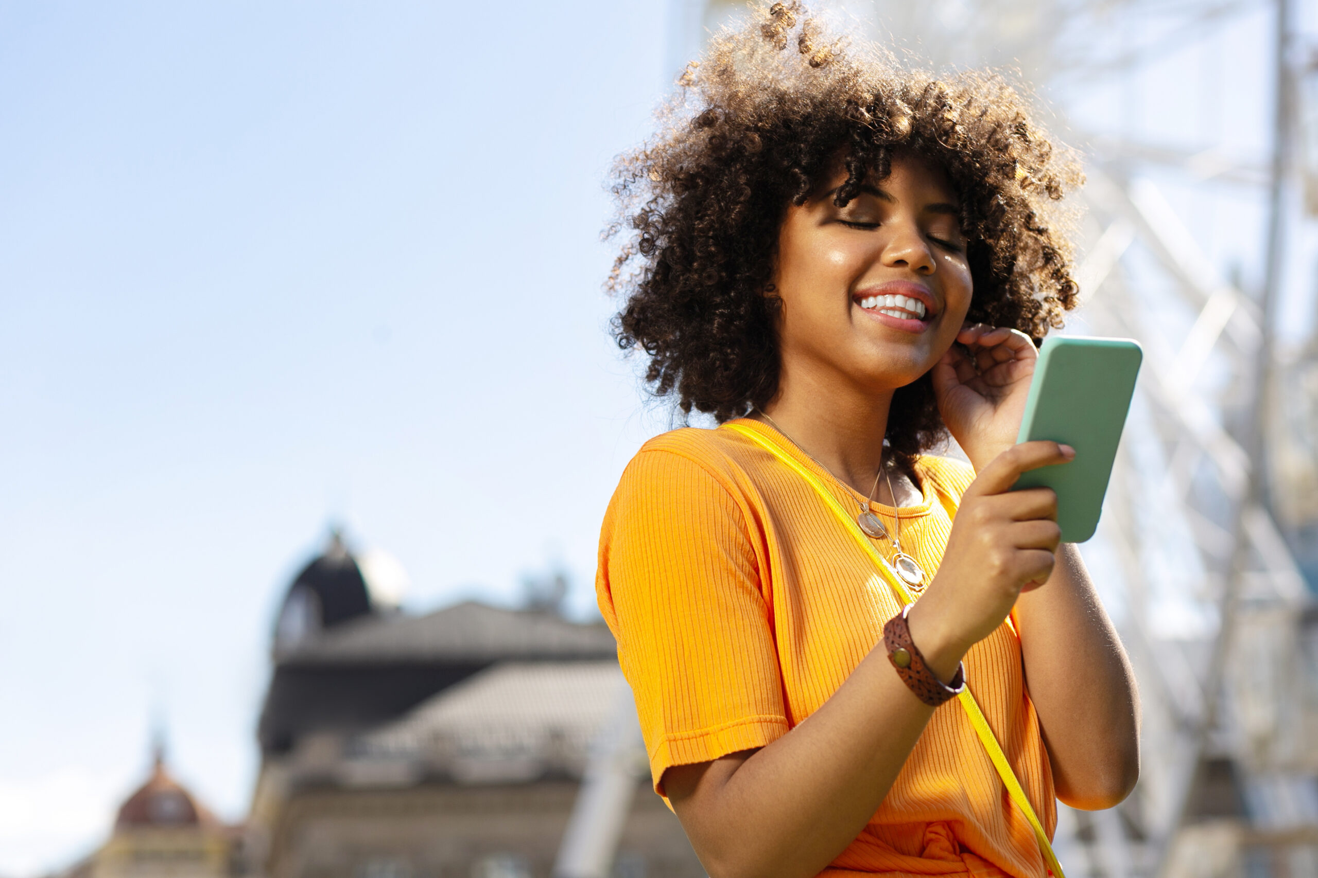 Heartwarming texts. Charming curly-haired woman reading text messages on her phone and smiling while visiting tourist attractions in another city